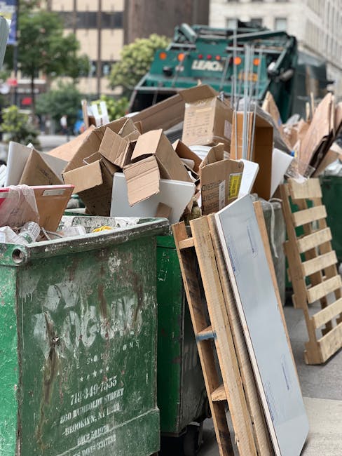 A large green waste disposal skip filled with a variety of packing materials, cardboard boxes, and other miscellaneous rubbish, situated on a city street with trees and buildings in the background. Th