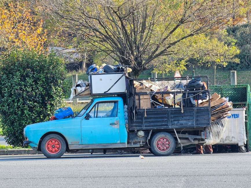 A blue flatbed truck parked on a street curbside, heavily loaded with various types of waste materials for rubbish removal, including cardboard boxes, black garbage bags, wooden pallets, and other hou