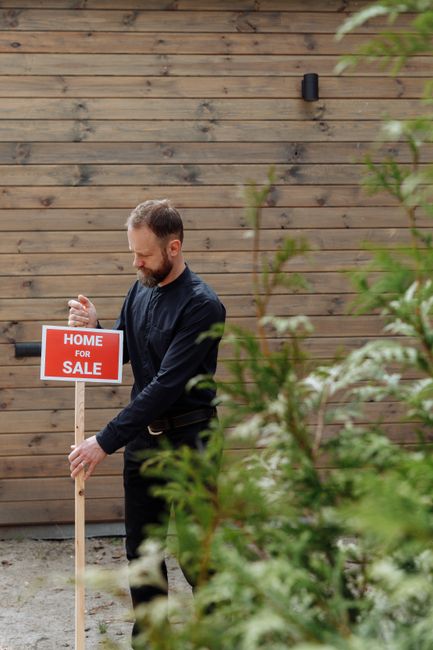 A man with a beard and dark clothing stands outdoors on a paved surface, facing a wooden fence with horizontal planks serving as a backdrop. He is holding a red and white 'Home for Sale' sign with bot